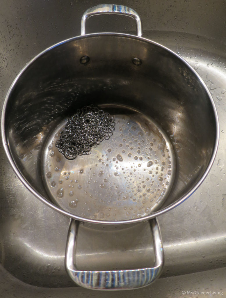photo of a pot while being scrubbed with a stainless steel chain mail scrubber, soap, and water. Chain mail scrubber and dirty bubbles are in the bottom of the pot.